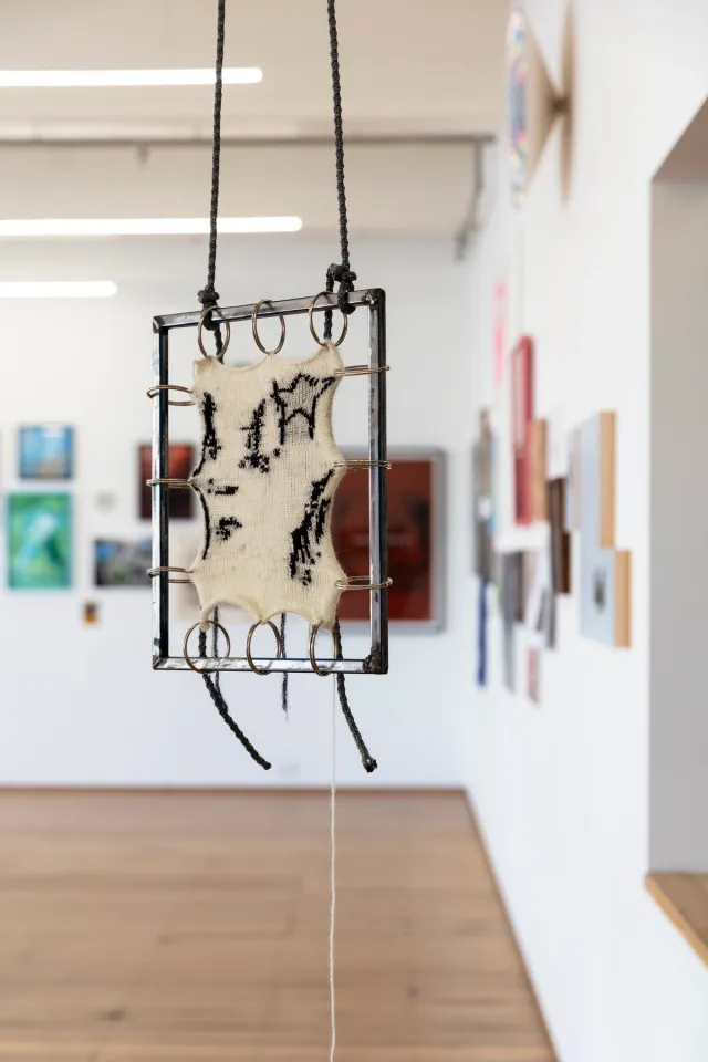 A small hanging sculpture made of a weaving stretched inside a metal frame suspended from large silver rings that loop around the metal frame and through the weaving. The weaving is white with abstract black marks woven into it. A black star is visible on the weaving. In the background, a salon hang is out of focus on the walls.