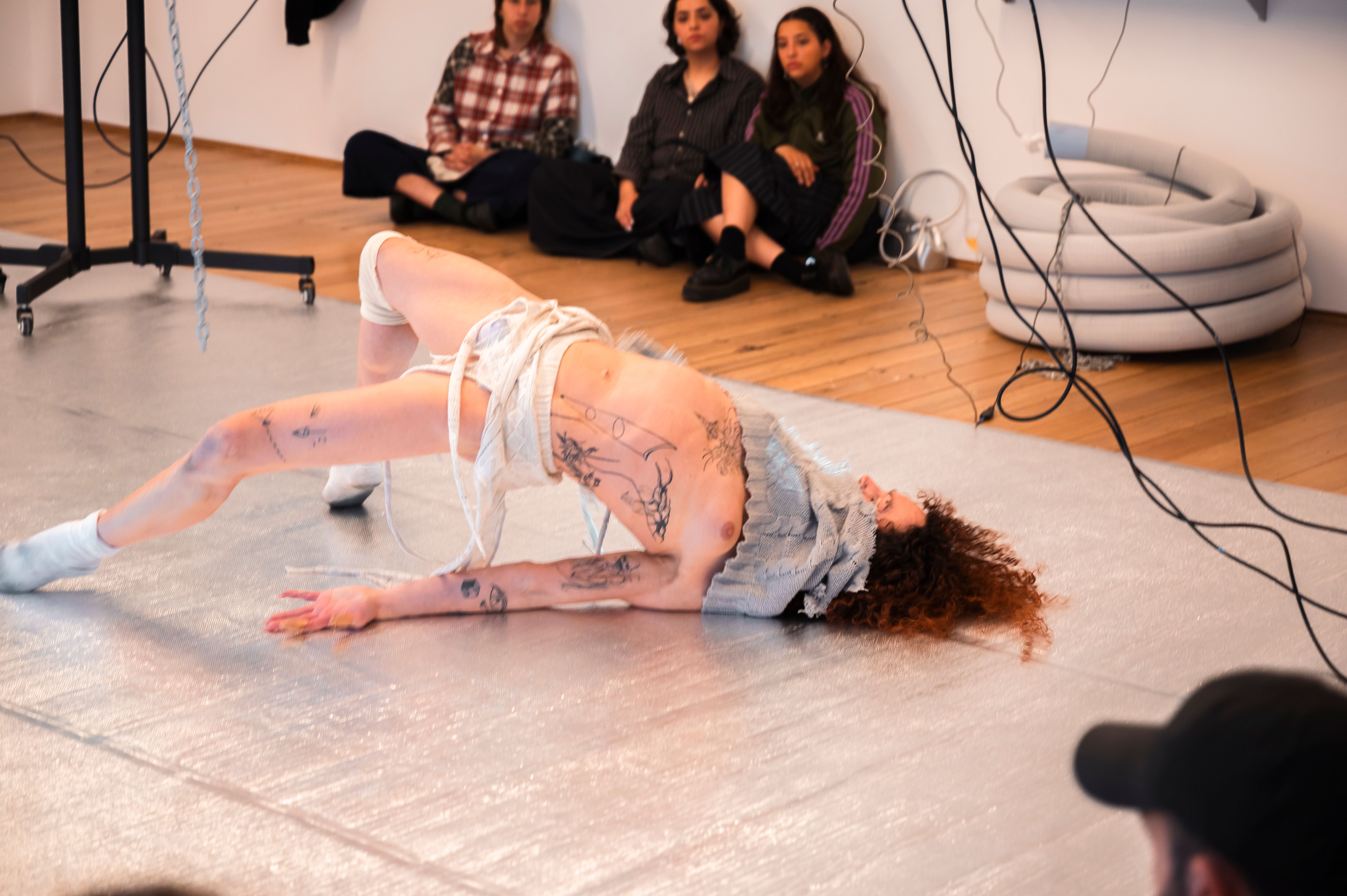 Andrea's body contorts in motion on the silver floor. There are audience members watching her in the top of the image.