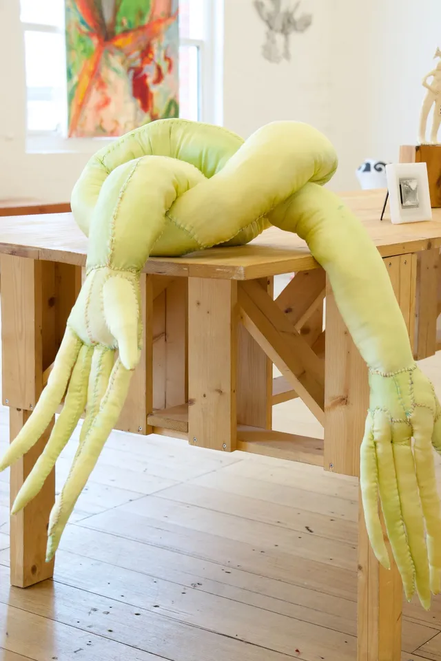 A light green stitched up fabric arm with elongated hands and fingers. The sculpture is tied into a knot and placed on the corner of a wooden table.