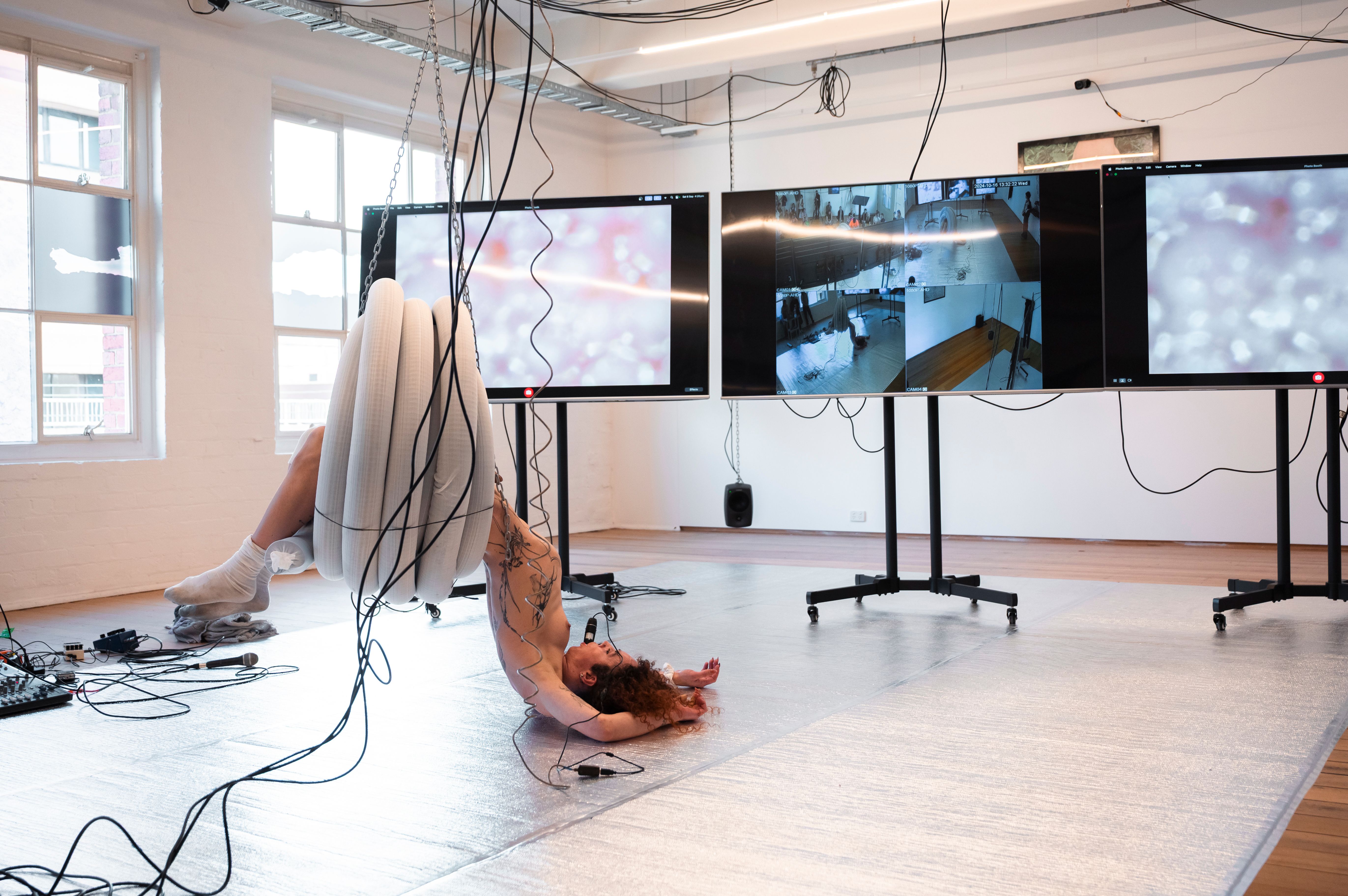 Andrea hangs from a coil of plastic pipe that is suspended from the ceiling with chains. She is flopped on to the silver floor in front of three large screens on wheels
