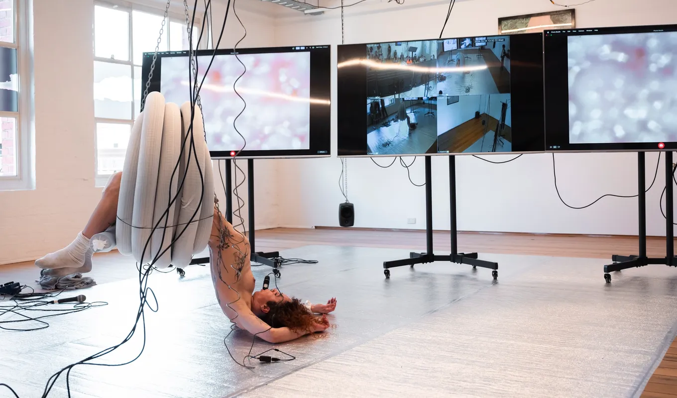 Andrea hangs from a coil of plastic pipe that is suspended from the ceiling with chains. She is flopped on to the silver floor in front of three large screens on wheels