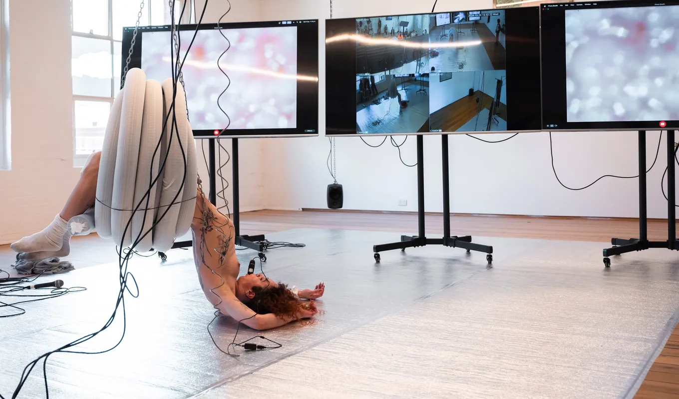 Andrea hangs from a coil of plastic pipe that is suspended from the ceiling with chains. She is flopped on to the silver floor in front of three large screens on wheels