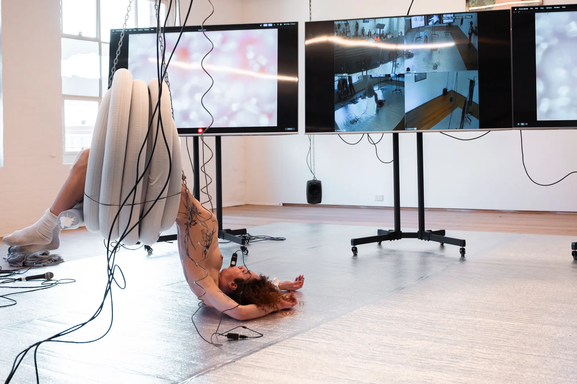 Andrea hangs from a coil of plastic pipe that is suspended from the ceiling with chains. She is flopped on to the silver floor in front of three large screens on wheels