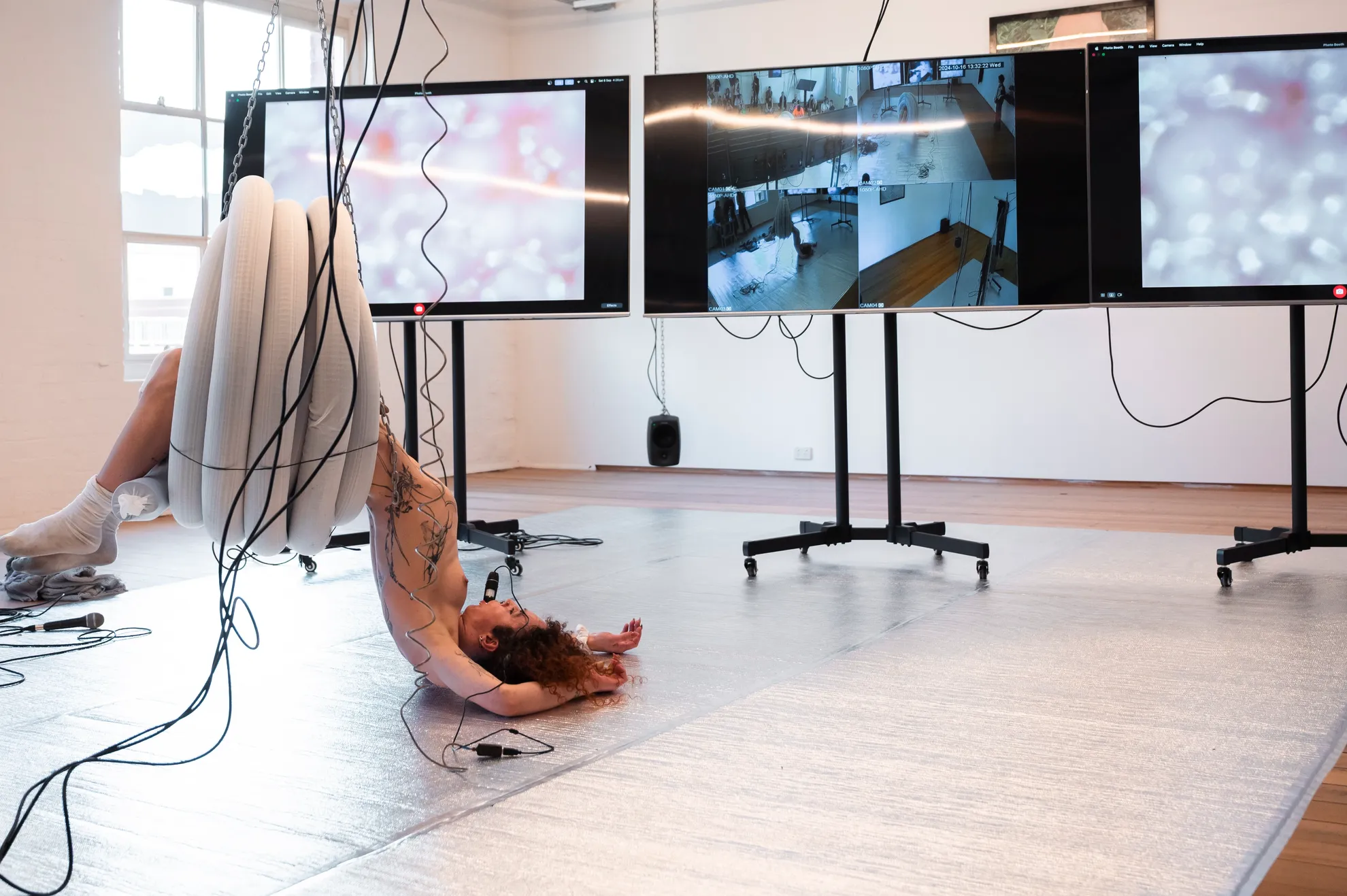 Andrea hangs from a coil of plastic pipe that is suspended from the ceiling with chains. She is flopped on to the silver floor in front of three large screens on wheels