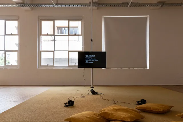 Installation view of 13 Years showing a screen suspended away from the gallery wall with two pairs of headphones laying on the floor connected to it. The screen is black with white text that reads "Peter Tosh tribute, Africa Connexions, Neville Legg and Patrick Jonathan, Radio Skid Row, Sydney 1987, 42:43 minutes".