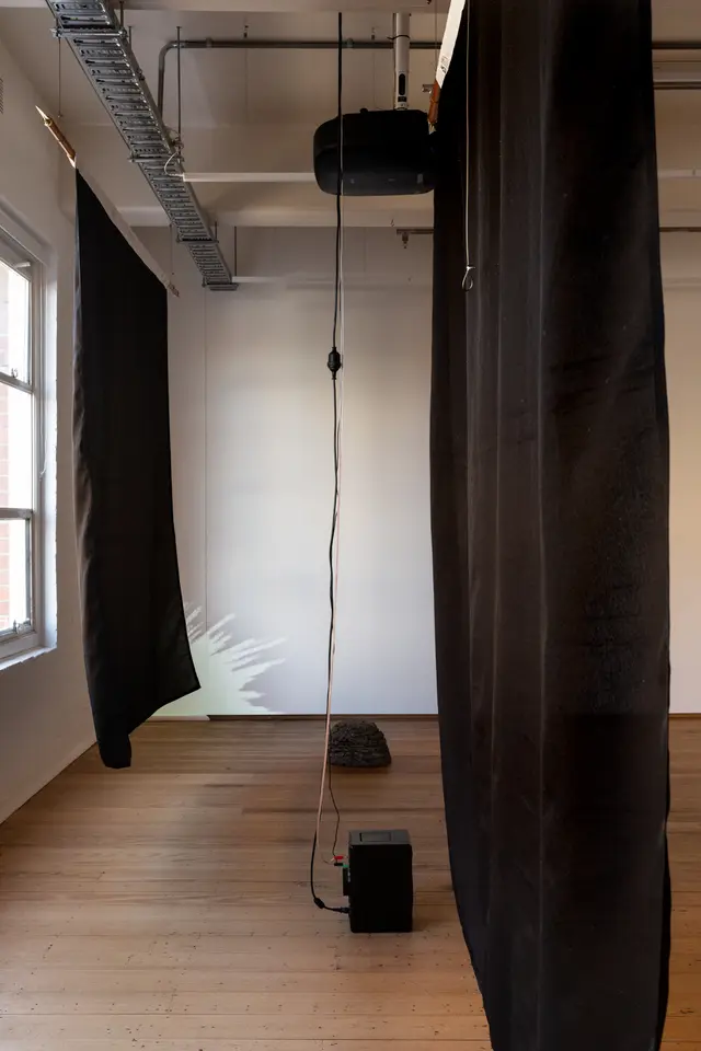 Installation view of "Big Pharmakon". The image is a side on view of two black semi-transparent sheets hanging from the roof in West Space Gallery. Between the two sheets a speaker sits on the floor with a plastic rock sitting behind it.