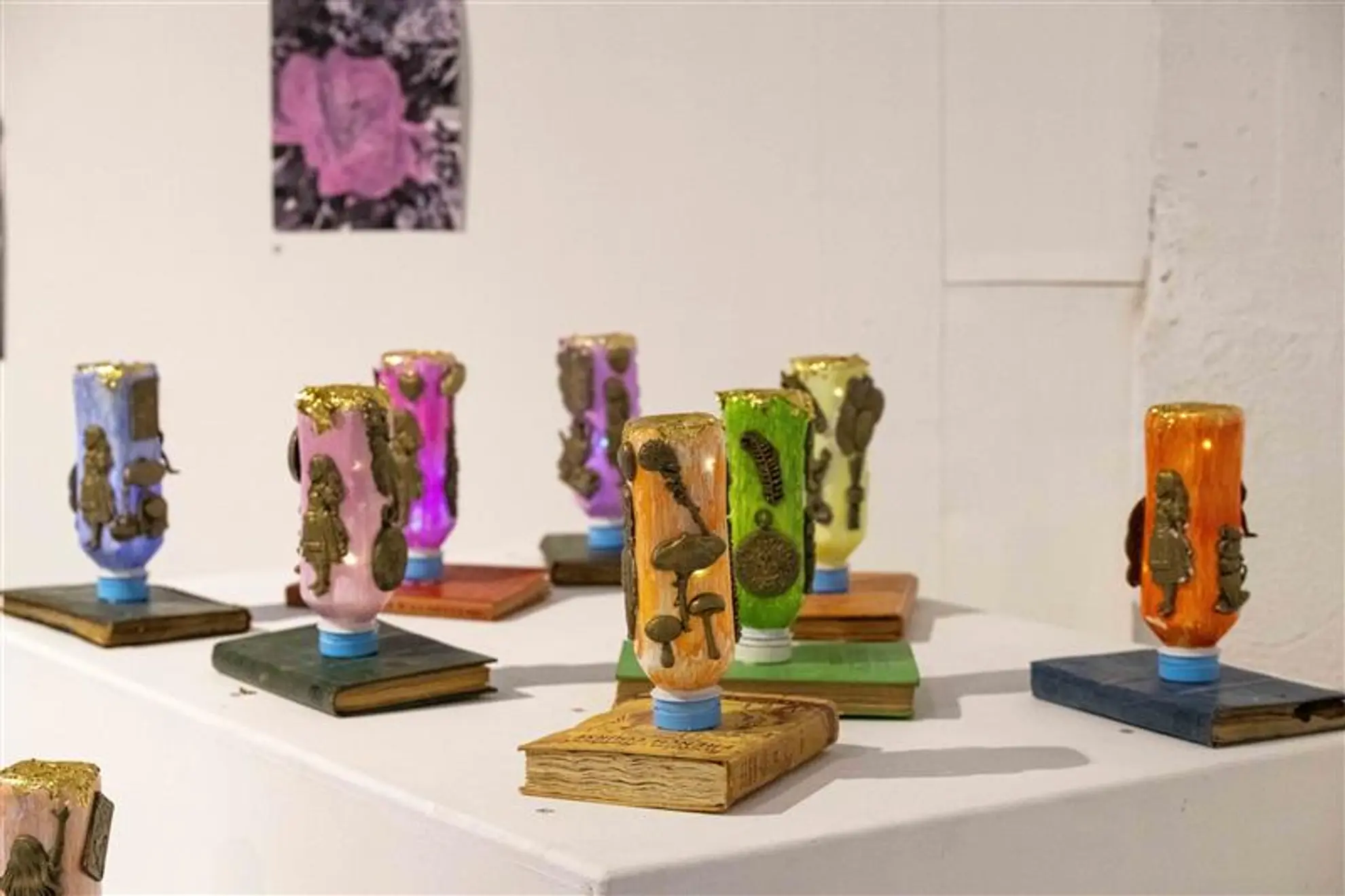 8 painted bottles stand upside down on a book. They have various bronze items stuck to them.