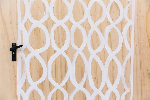 a close up of a white line painting of a screen door on plywood. There is a screen door handle that has been attached to the wooden board.