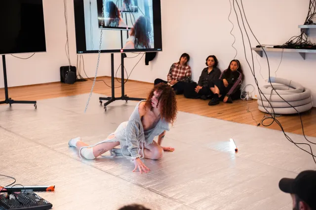 Andrea leans sitting on the silver floor of the gallery looking back over her shoulder. Three members of the audience sit next to a large screen watching her perform.