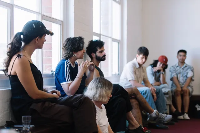 A group of audience members seated along a wooden bench in front of two nine grid windows.