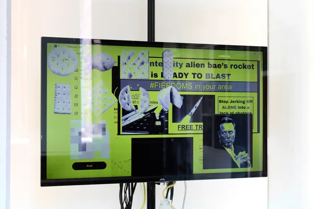 Behind glass, a landscape screen in a black frame displays black-and-white images of biscuits, a rocket, and Elon Musk, with partially obscured text.