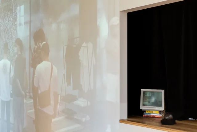 Installation view of "Big Pharmakon". On the left of the image a small analog television sits on a countertop on a pile of books. A black curtain hangs behind the counter. To the right of the counter is a large projection of a group of people standing around.