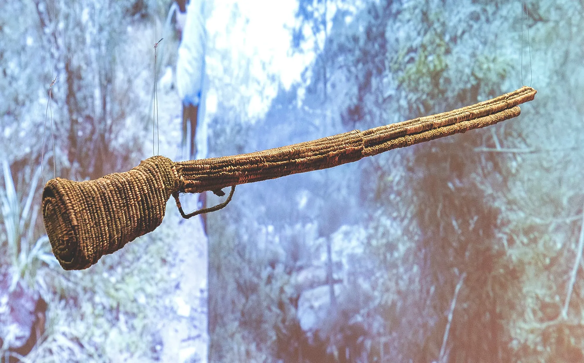 A gun made through some densely, thick, weaved natural kind of branch or material, suspended lightly with a very thin wire in-front of a large projection of a rustic, hazy landscape of bush.