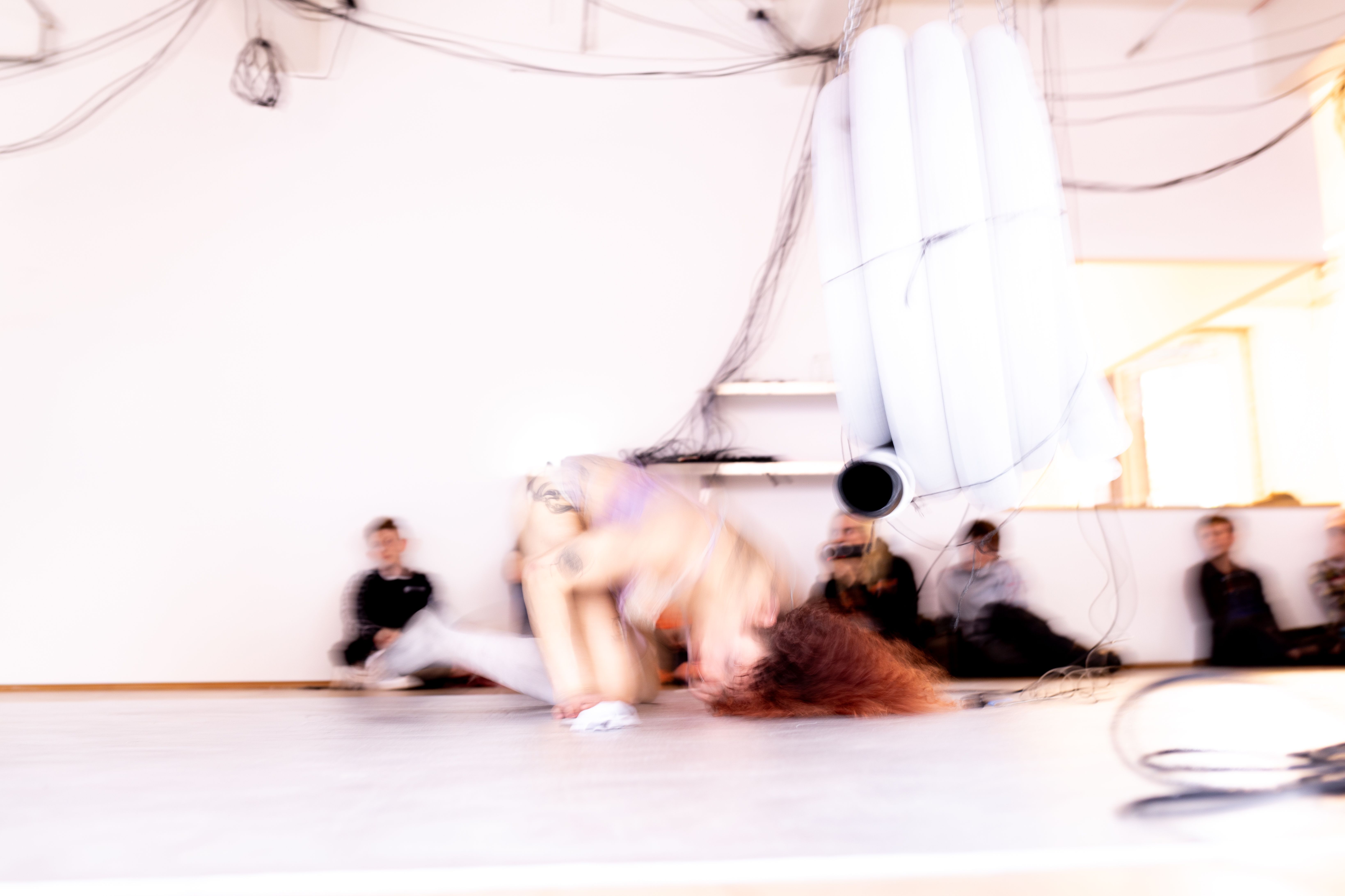 Blurry image of Andrea Illés hunched over the floor in front of an audience seated on the floor.