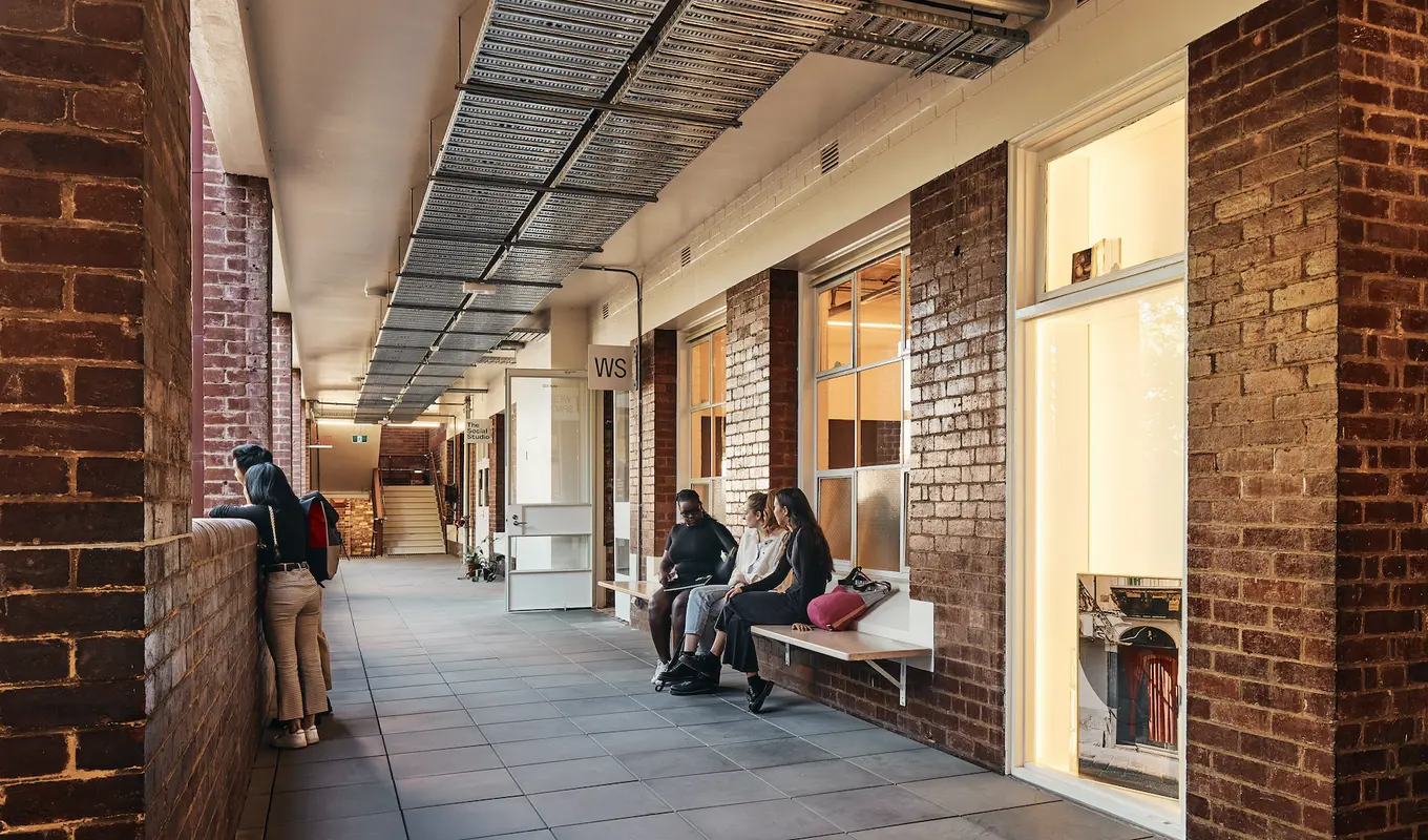 A walkway with people sitting on a bench against the facade of West Space, and other people looking out over the balcony. In the middle of the image you can see the open door to West Space.