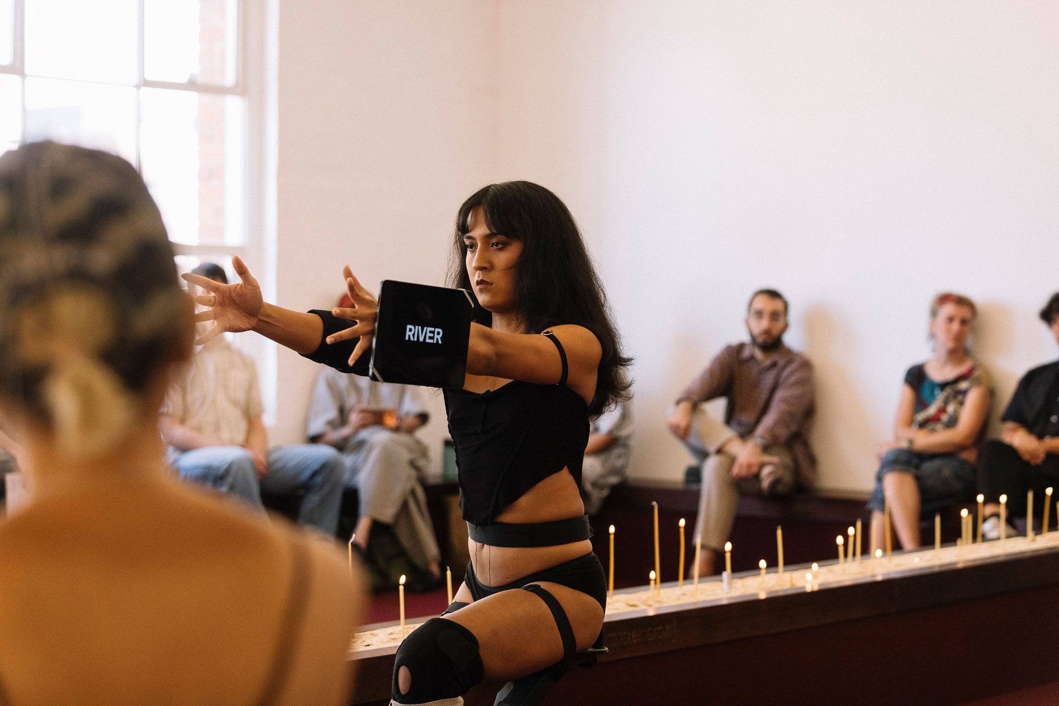 Ari performing in front of a row of lit candles. She has an ipad attached to her wrist that has text on the screen that says "River".