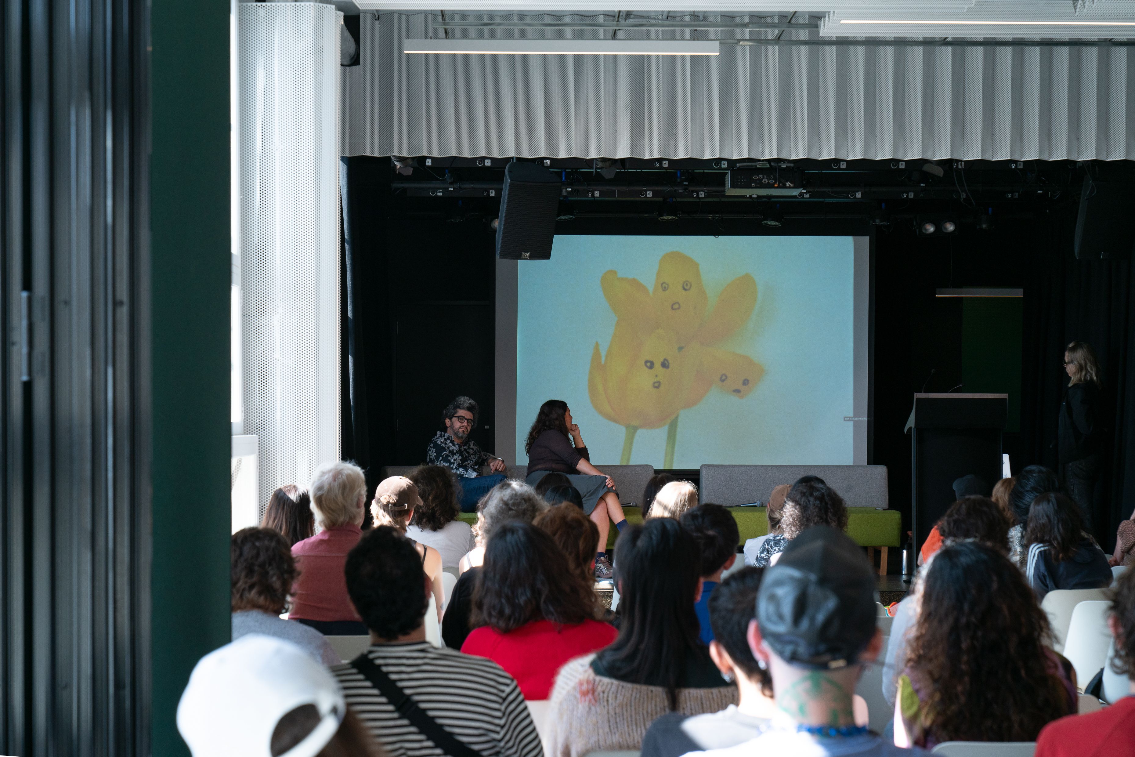People watching a screen in a room, which displays two yellow tulips with three faces on the petals against a white background..