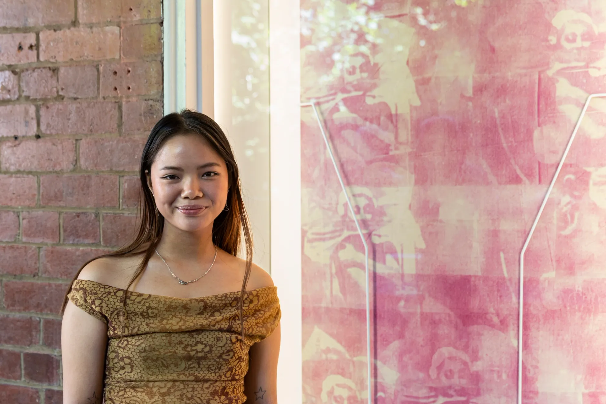 A smiling woman stands in front of her artwork on display in the West Space window. The artwork is a pink faded ink transfer of a woman in many scenes. The artist is smiling and looking at the camera