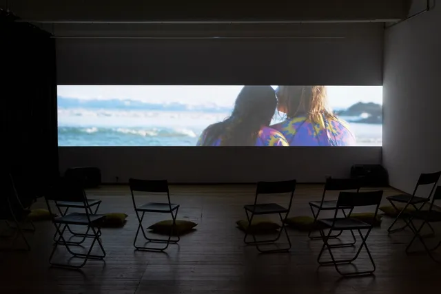 Video install playing in a space with empty chairs and cushions.