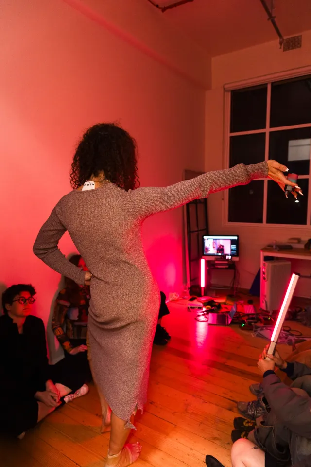 A figure wearing a grey woolen dress poses with one hand on her hip and one hand outstretched in front of a room lined with people in red lights