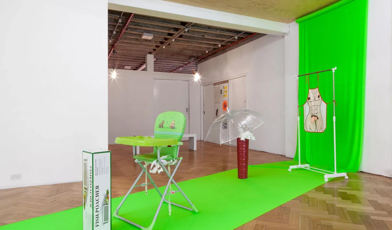 A floor to ceiling draped piece of bright green fabric with various objects placed on top including a baby chair, a clear opened umbrella, and a clothing rack displaying an apron with a human body illustration with a penis sculpture.