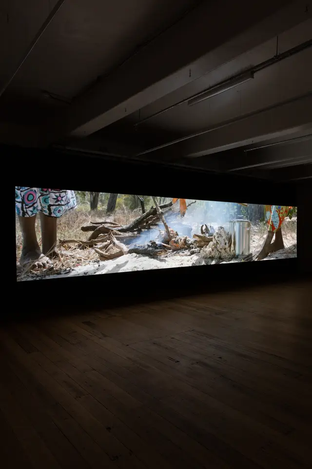 A long rectangular film still is projected on the gallery wall. The image shows smoke coming from a fire outside and a persons legs standing next to it.