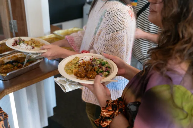 A guest holds a plate of Persian food, their face cut out of frame with the camera centred in on the plate. Another guest in the background is serving themselves another plate of Persian food.