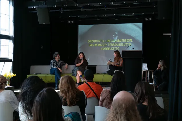 Three individuals sit on a green couch on a stage discussing with one another. A projection screen is visible behind them. A crowd of people are seated on white chairs listening.