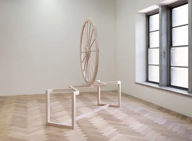 Side view of a large wooden sculpture or a wheel placed on a bench. The wheel is balancing on the intersection of two pieces of wood crossing over on the bench. The wheel is placed in front of a large window with natural light shinning through.