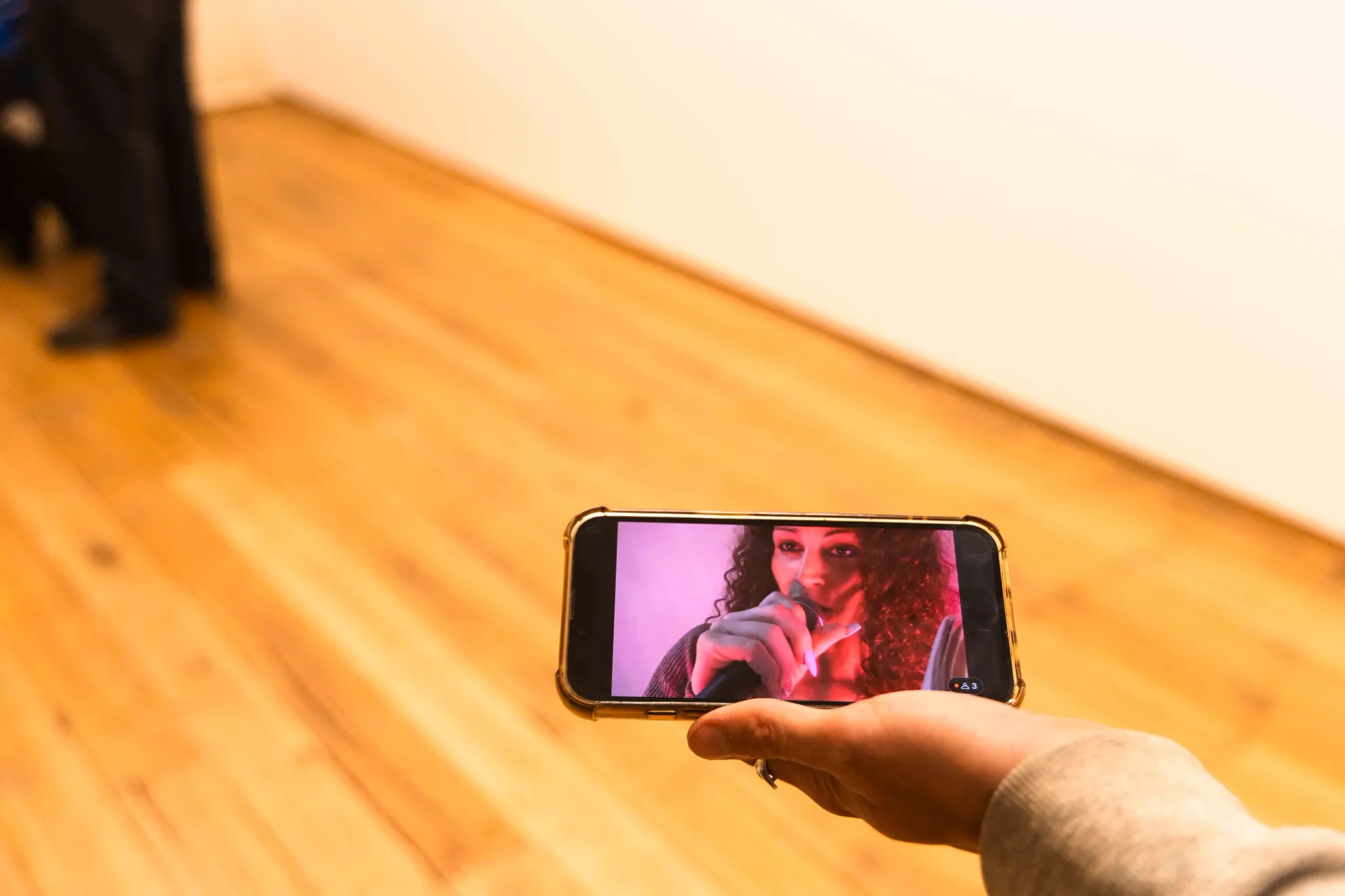 A hand outstretched holds an iphone watching a livestream performance. On the phone screen we see a woman holding a microphone with long curly hair.