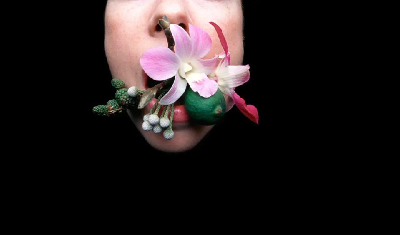 A persons face sticking out of a black background with their mouth full of flowers.
