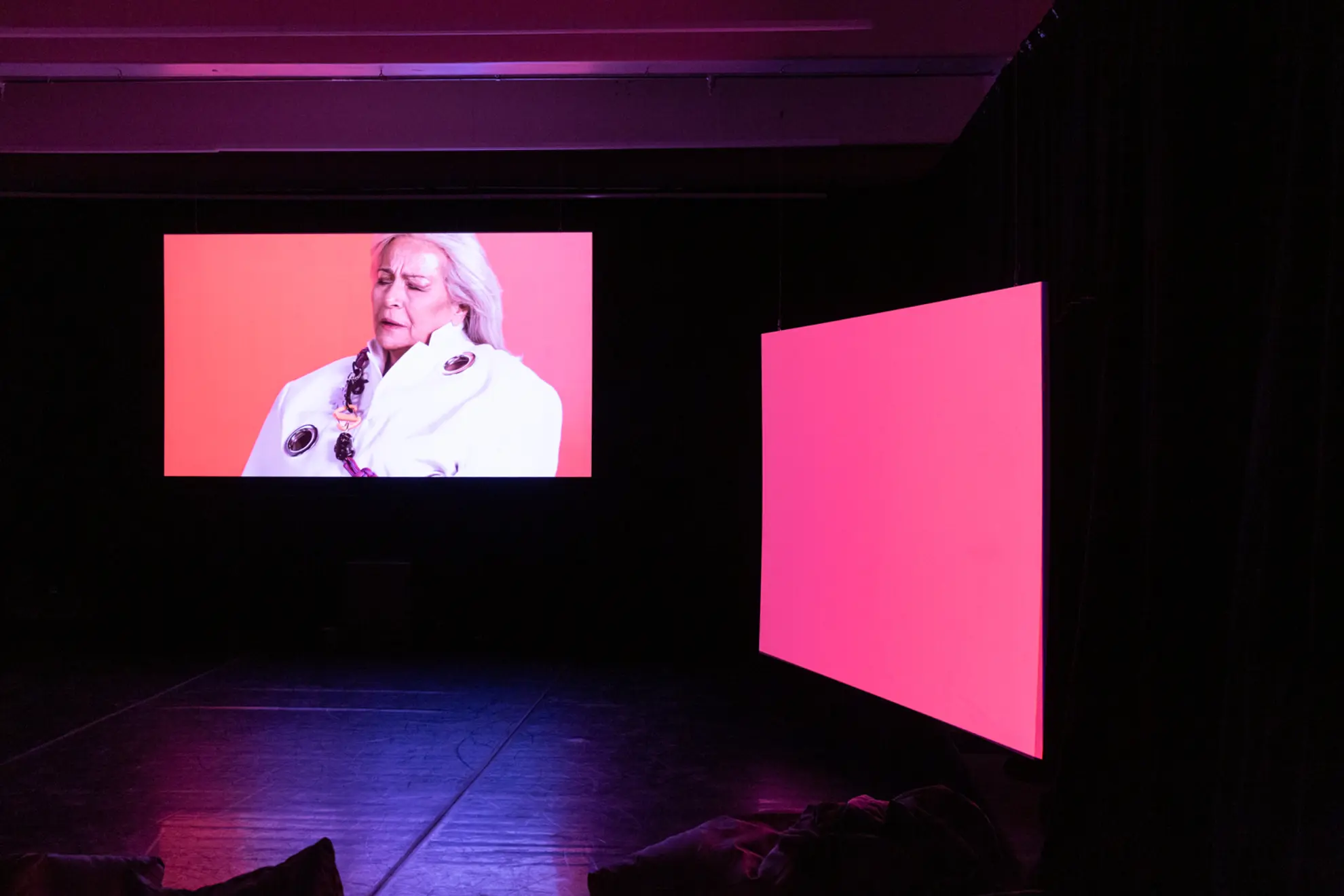 A photograph of artist HOSSEI's audio-visual artwork THUNDERBLOOM. Three screens glow with the projected images of performers, bright backgorund colours and captioned lyrics.