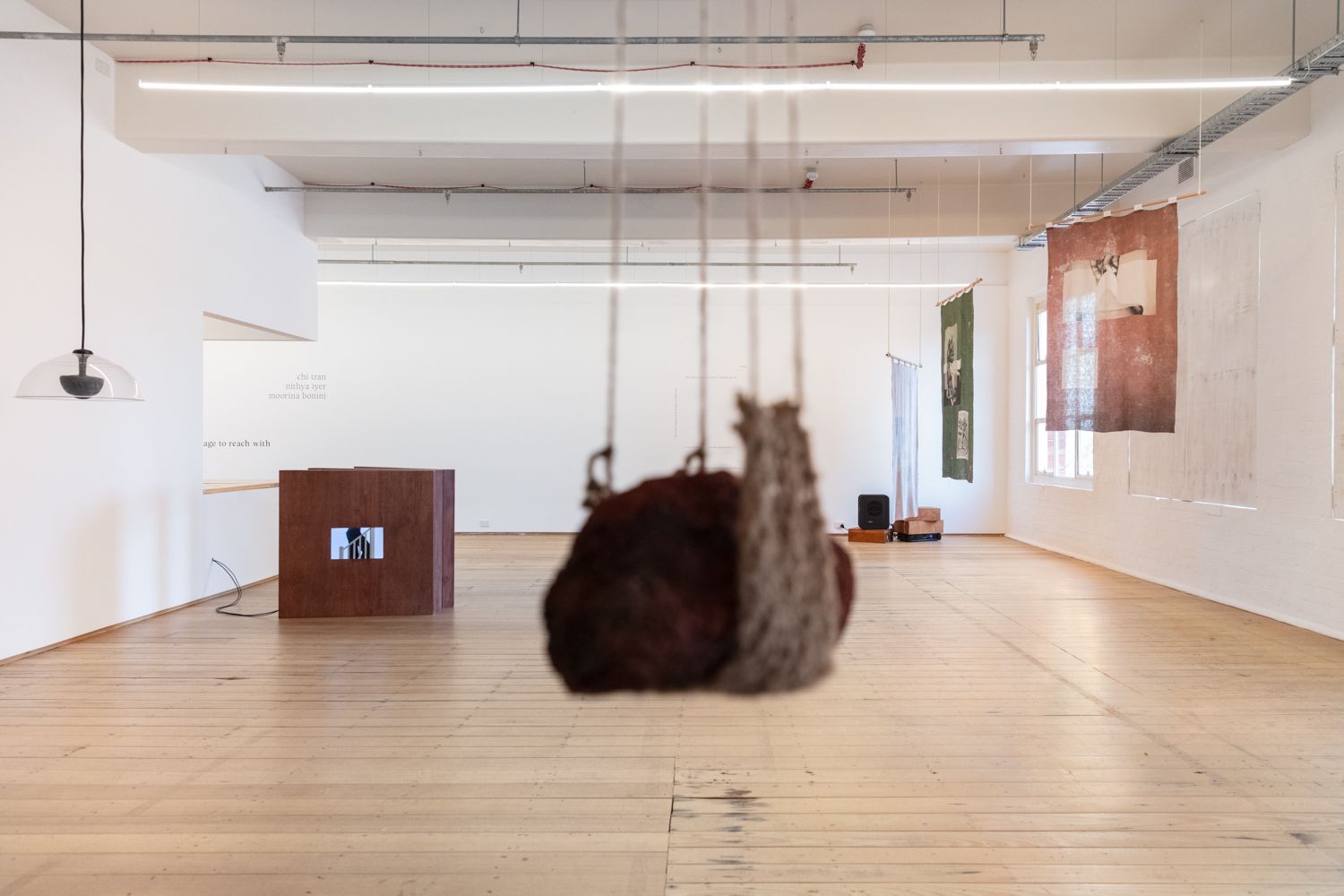 Install view of 'language to reach with' shot from behind Moorina Bonini's piece of hanging earth in blur with the focus on the rest of the gallery space.