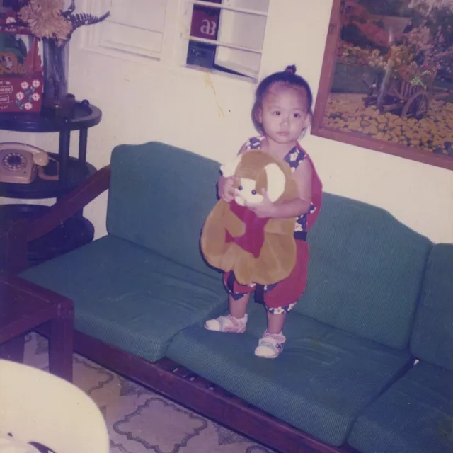 A photograph of a young girl holding a soft toy standing on a green couch in a living room.