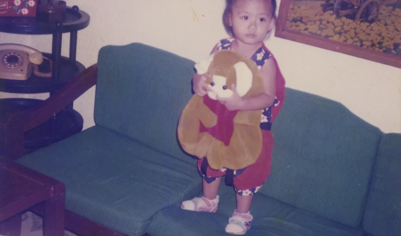 A photograph of a young girl holding a soft toy standing on a green couch in a living room.