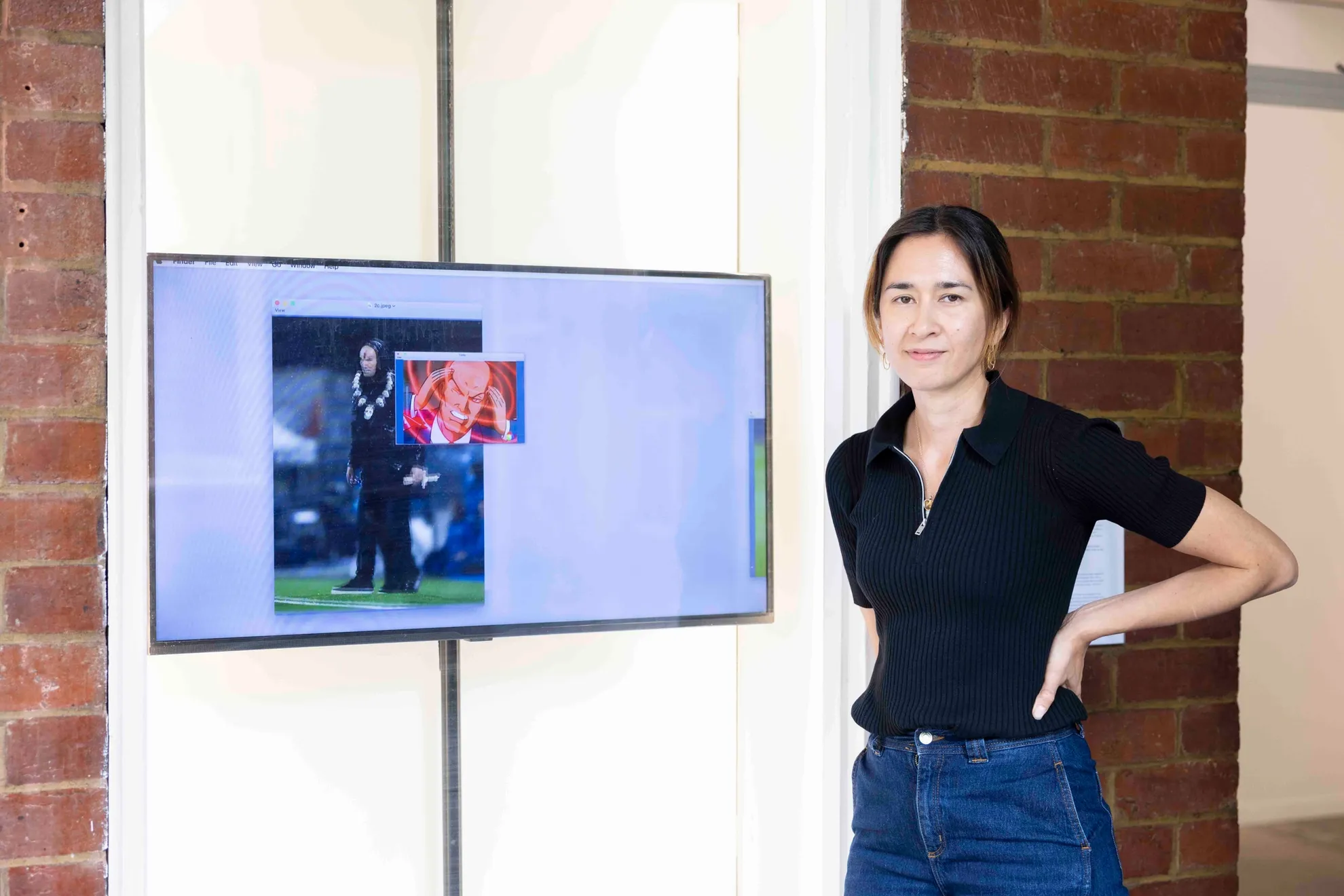 The artist poses next to a projection work on a screen inside of a tall narrow window. Their hands are on their hips smiling subtly.