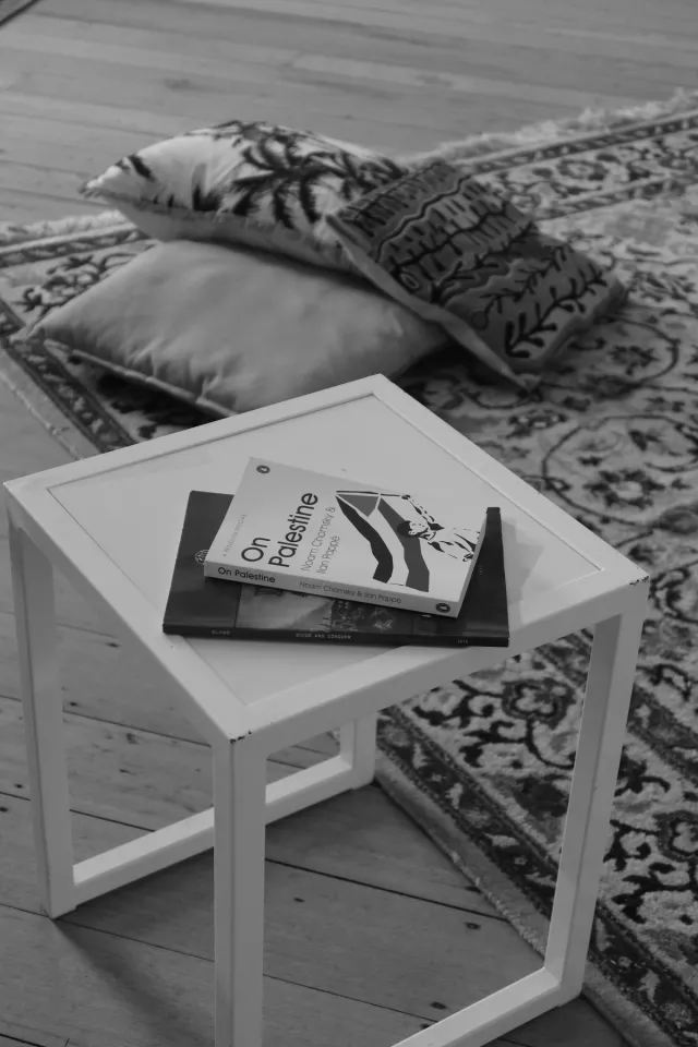 A black and white image of the 'On Palestine' Book on top of a second book on a square angular side table. The background displays a patterned rug and a stack of three patterned pillows.