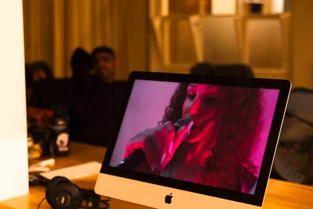 A mac computer screen sits in the west space office with a figure holding a microphone is close up on the screen. She has long white nails and long dark curly hair