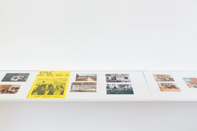 The image shows a white shelf suspended off the wall with a series of images placed on it. There are a number of colour photographs showing groups of people, musicians and protest posters. There is a yellow newspaper clipping with the title PAC.