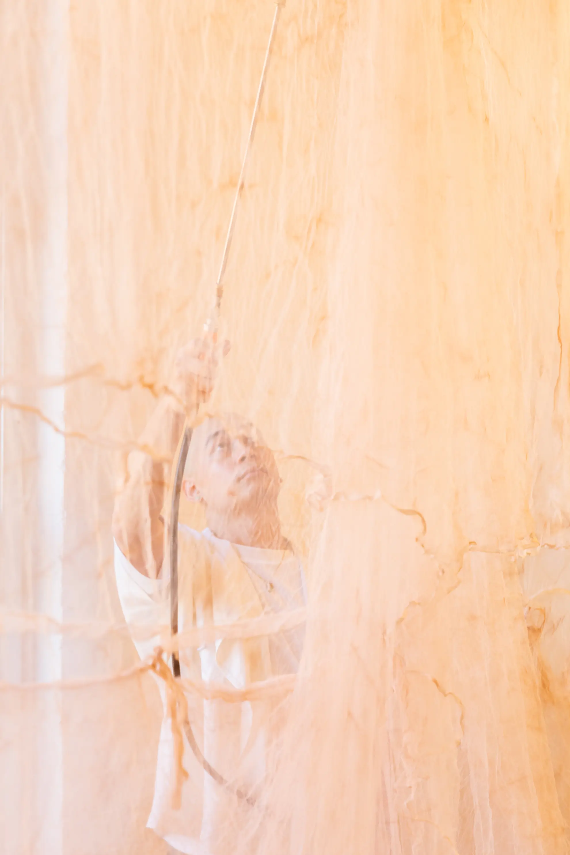 A subtle, muted image of the artist, Ngo, behind the died mosquito nets in the exhibition. They are spraying the nets with a thin, short hose attached to a medium metalic can. Ngo stares upward, intensely at the net. The image is of a beautiful haze.