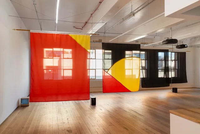 Installation view of "Big Pharmakon". The image shows 4 semi-transparent fabric sheets in front of 3 windows in the West Space Gallery. The first sheet is red with a small triangular yellow section in the top right. The second shows a segment of a yellow circle bisected by a red triangle in the bottom left over a black background. The third and fourth sheets are completely black. On the ground in front of the sheets are two speakers and a small analog television.