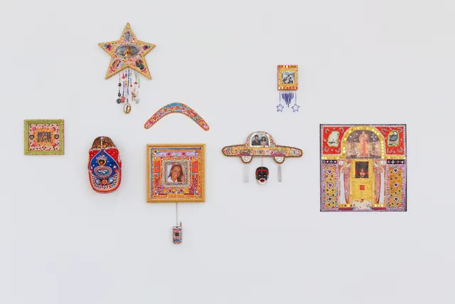 A close-up image of Aretha Brown's eight embellished wall assemblages hung in an uneven cluster. From left to right, the first work is a framed image of Beyonce, surrounded by many small colourful and reflective gems. The second is a 'Western Bulldogs' football cap, also embellished with jewels, primarily red and blue. Above those two is a jewelled star frame, with 'Kath' from 'Kath and Kim' wearing a t-shirt with an indigenous flag. There are jewelled tassells hanging off the star. Next is a small boomerang embellished with pink, blue and red gems. Below that is an image of Mariah Carey in a square jewelled frame, with a jewelled flip-phone attached with a chain. The next is a cartoon car filled with gems, with an image of two people and a face hanging below. Above is a very small square jewelled frame, with blue beaded stars hanging off it. Finally on the right side is a large square frame, with a photograph of Robert Smith, two small photos of birds and brightly coloured gems.