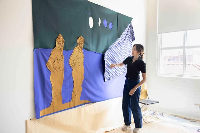 Artist Tintin Cooper stands in front of a large textile artwork in her West Space Studio. The artwork is of 2 large figures sewn in standing in front of a background that is half blue and half dark green with a central horizon line. There are 4 circular shapes at the top of the artwork