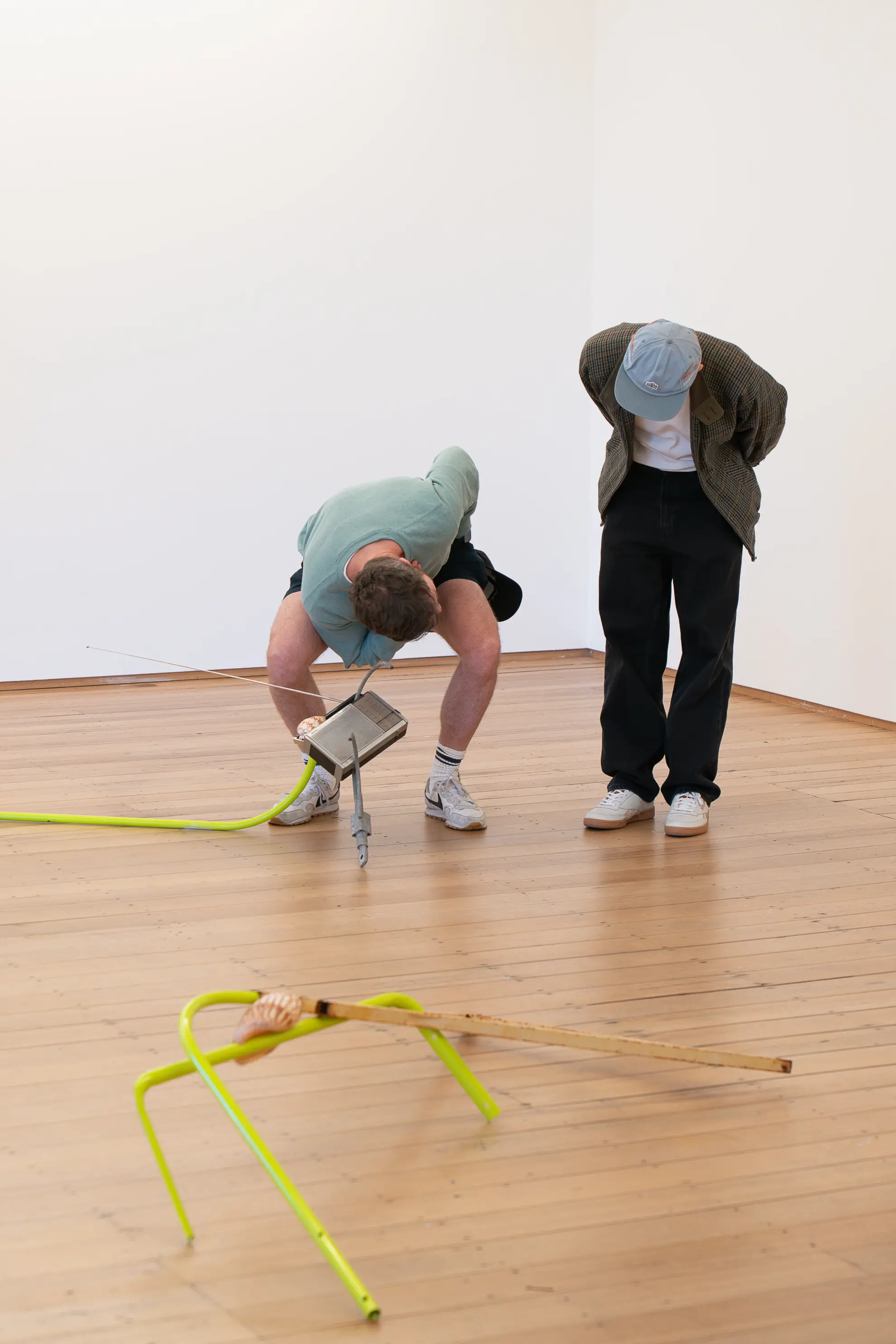 Two gallery visitors are bent down listening to a radio on the floor attached to a metal pole. A second sculptural install is in the forefront, containing metal, wood and a shell.