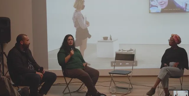 Three people in conversation on the chairs with microphones in front of a video projection on the wall.
