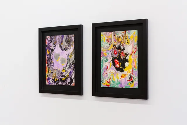 a pair of two 'marbled' works on paper. The works are both in black frames on a white wall. The works are bright colours and contain abstracted shapes eluding to scary eyes and faces.