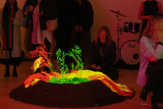 In a softly lit room, several people gather around a vibrant installation of plants growing from a mound of soil, lit in green, red, and yellow hues. One person sits near the display with their eyes closed, while a drum set sits in the background.