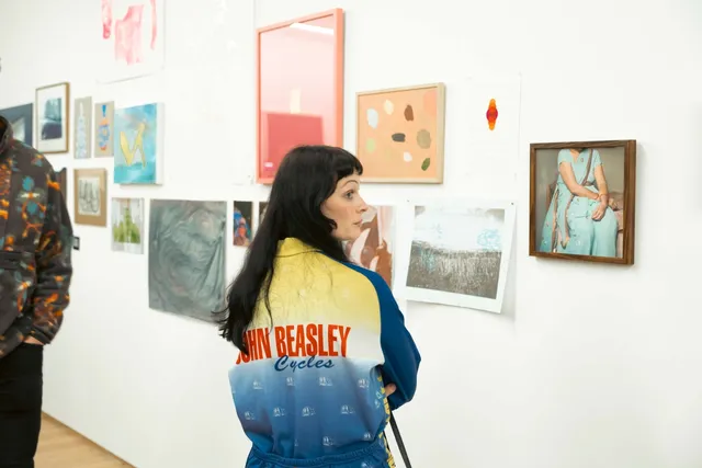 a woman with long dark hair and fringe stands side on to a wall full of artworks on show for the West Space fundraiser. She is wearing a yellow and blue jacket with red text that says "John Beasley Cycles"