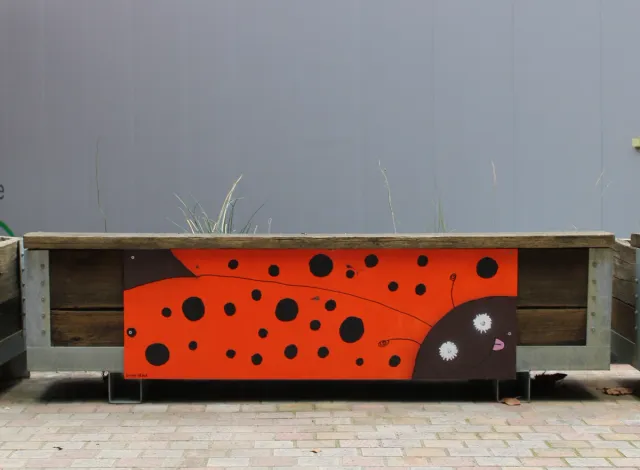 A rectangular board painted into a ladybug design thats been drilled onto the side of a wooden plant crate.
