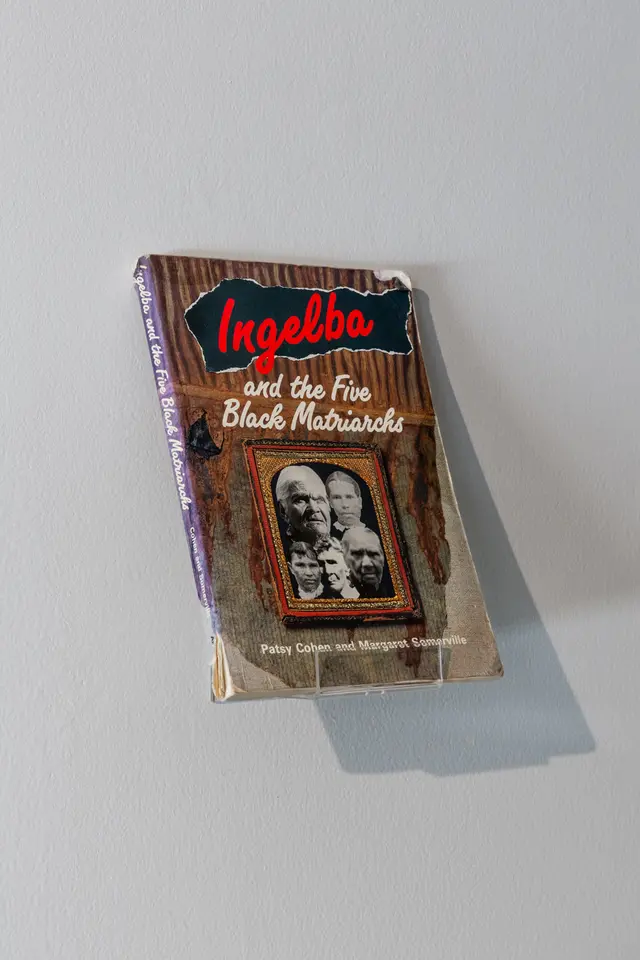 A book leans up on a acrylic stand against a white wall, its shadow casting behind and below it. The book sit at an angle, and is titled "Ingelba and the Five Black Matriarchs"